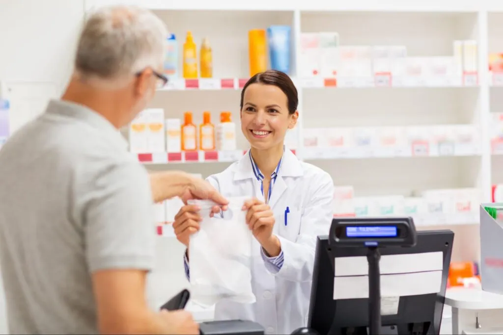A smiling pharmacist in a white coat hands a prescription bag to a customer at a pharmacy counter.