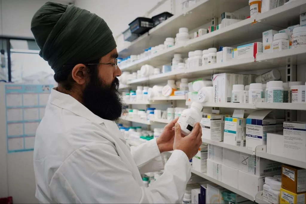 A pharmacist in a white coat is looking at a medication bottle, in front of a fully stocked shelf.