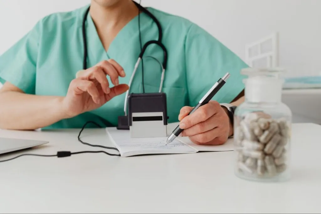 A healthcare professional wearing green scrubs and a stethoscope sits at a desk, writing in a notepad with a pen in one hand and preparing a self-inking stamp with the other, with an unlabeled jar of capsules in the front.