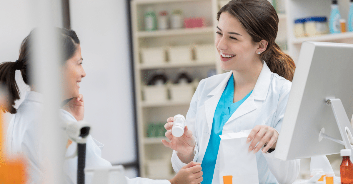 Smiling pharmacists handing a prescription bottle and bag to a customer at the pharmacy counter.