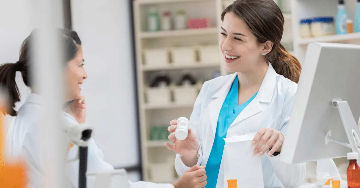 Smiling pharmacists handing a prescription bottle and bag to a customer at the pharmacy counter.