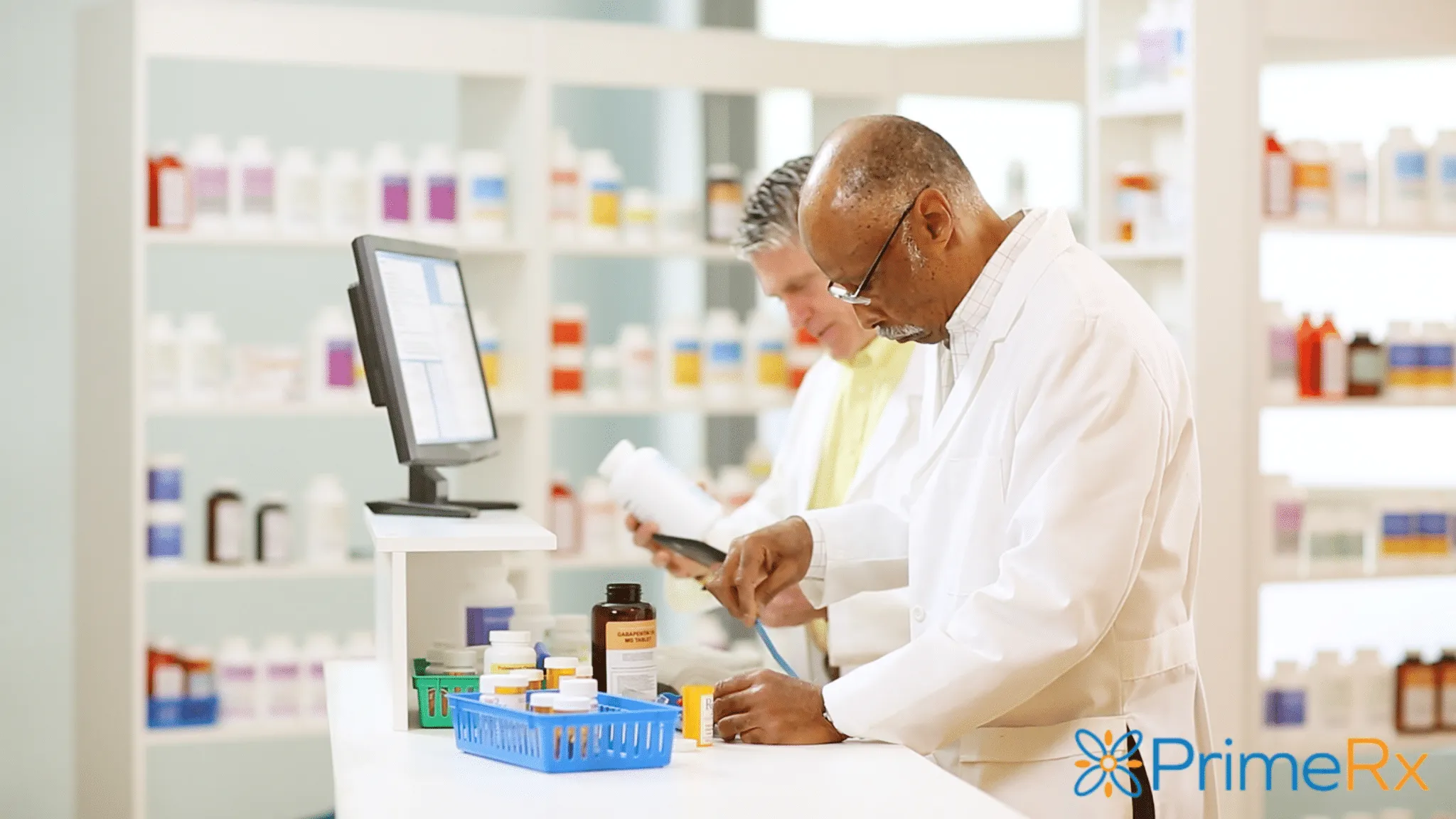 Two pharmacists are working behind the counter in a pharmacy, filling prescriptions near a computer with the PrimeRx logo.