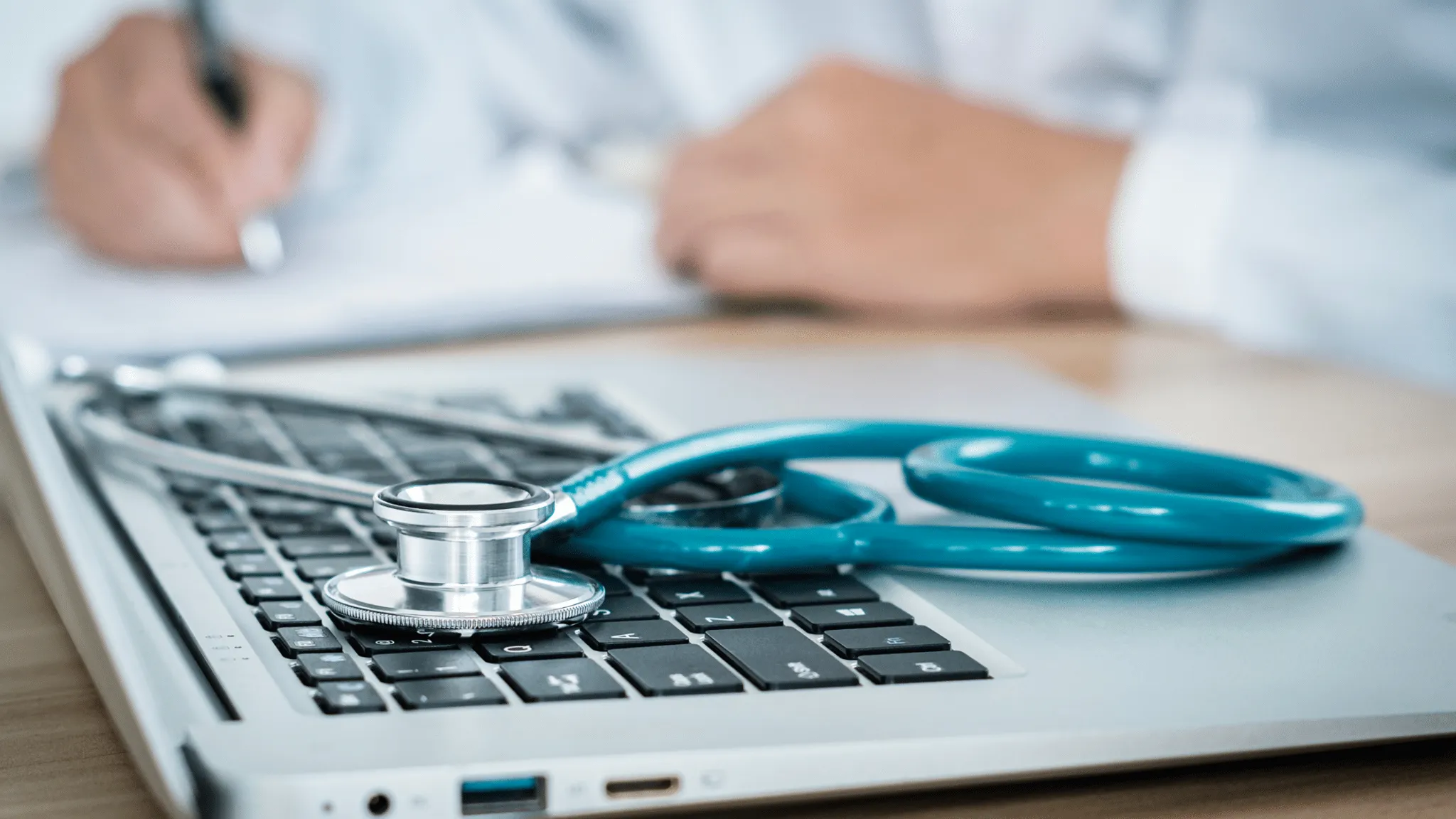 A stethoscope on a laptop with a doctor writing in the background.
