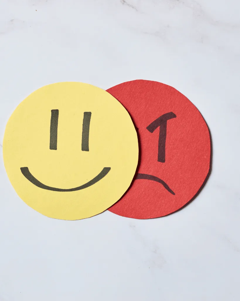 A yellow paper smiley face and red paper angry face against a white surface.
