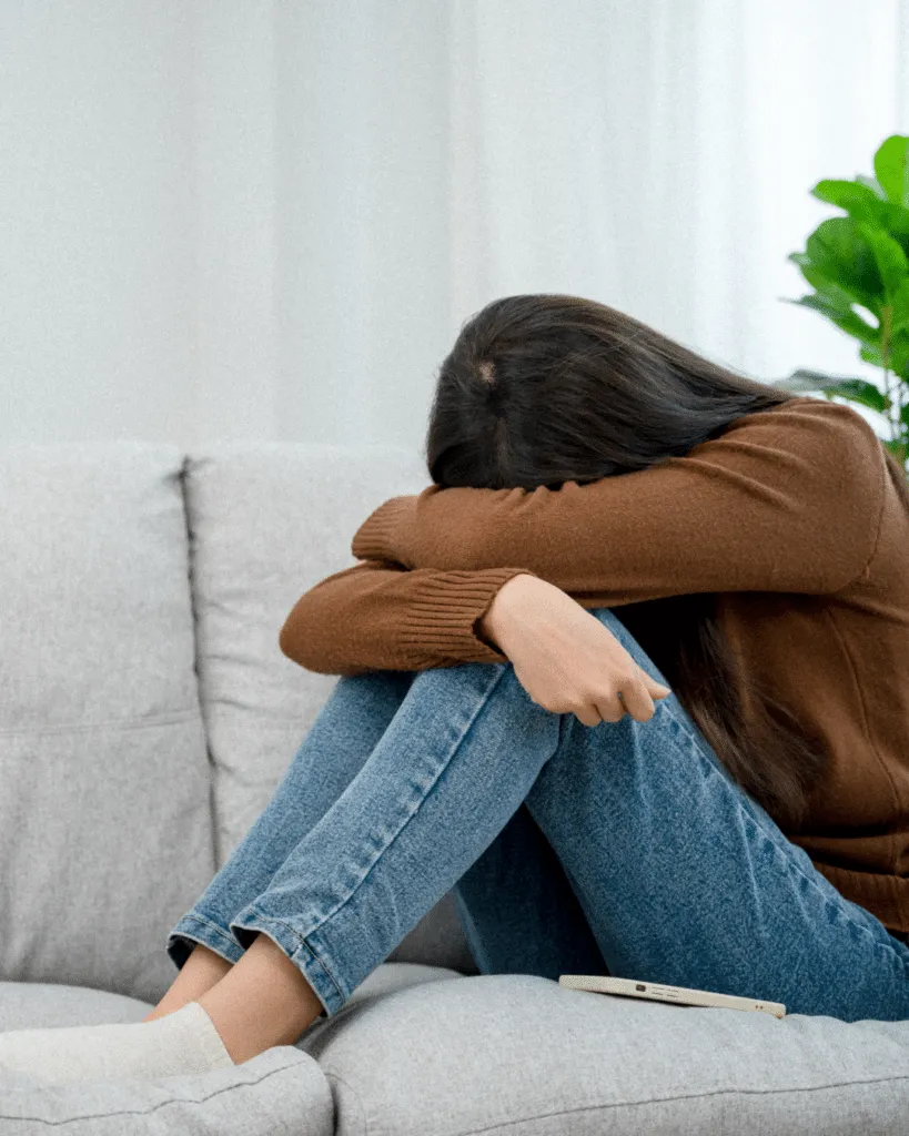 A woman sitting on a couch with her head resting on her arms, looking distressed, with a smartphone beside her.