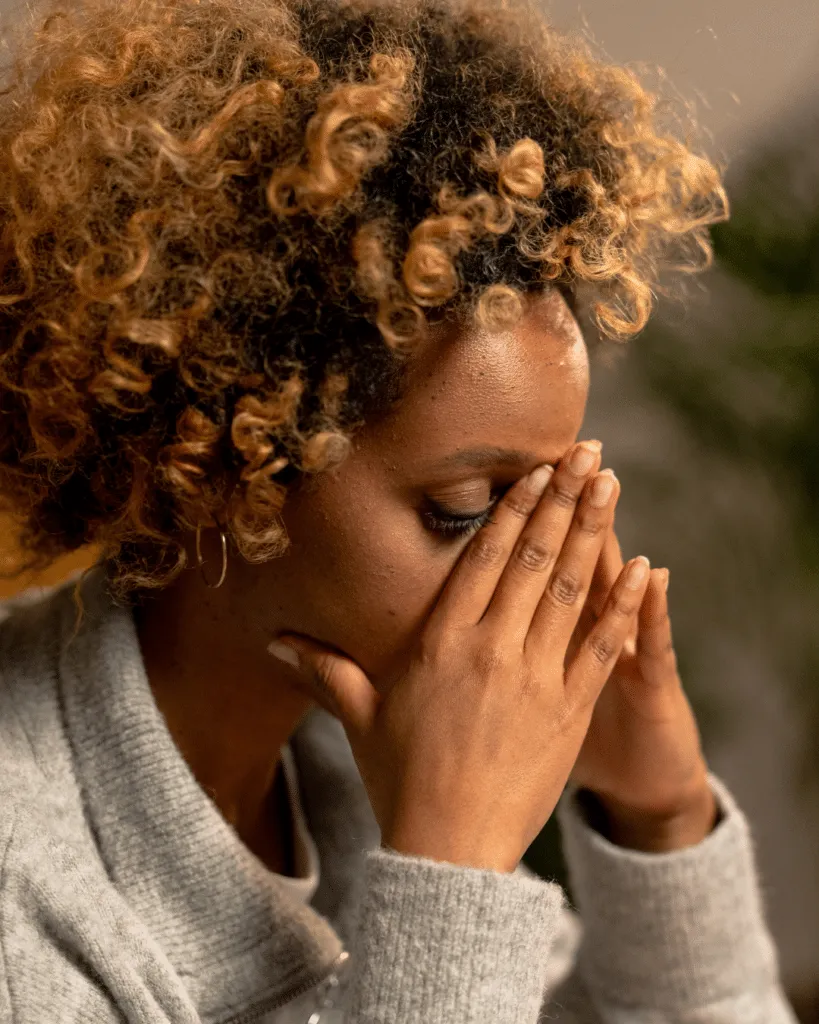 A woman holding her head in her hands, wearing a gray sweater against a neutral background.
