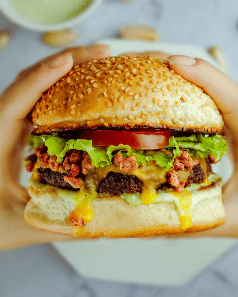 Close-up of hands holding a large sesame seed burger with lettuce, tomato, melted cheese, and a beef patty.
