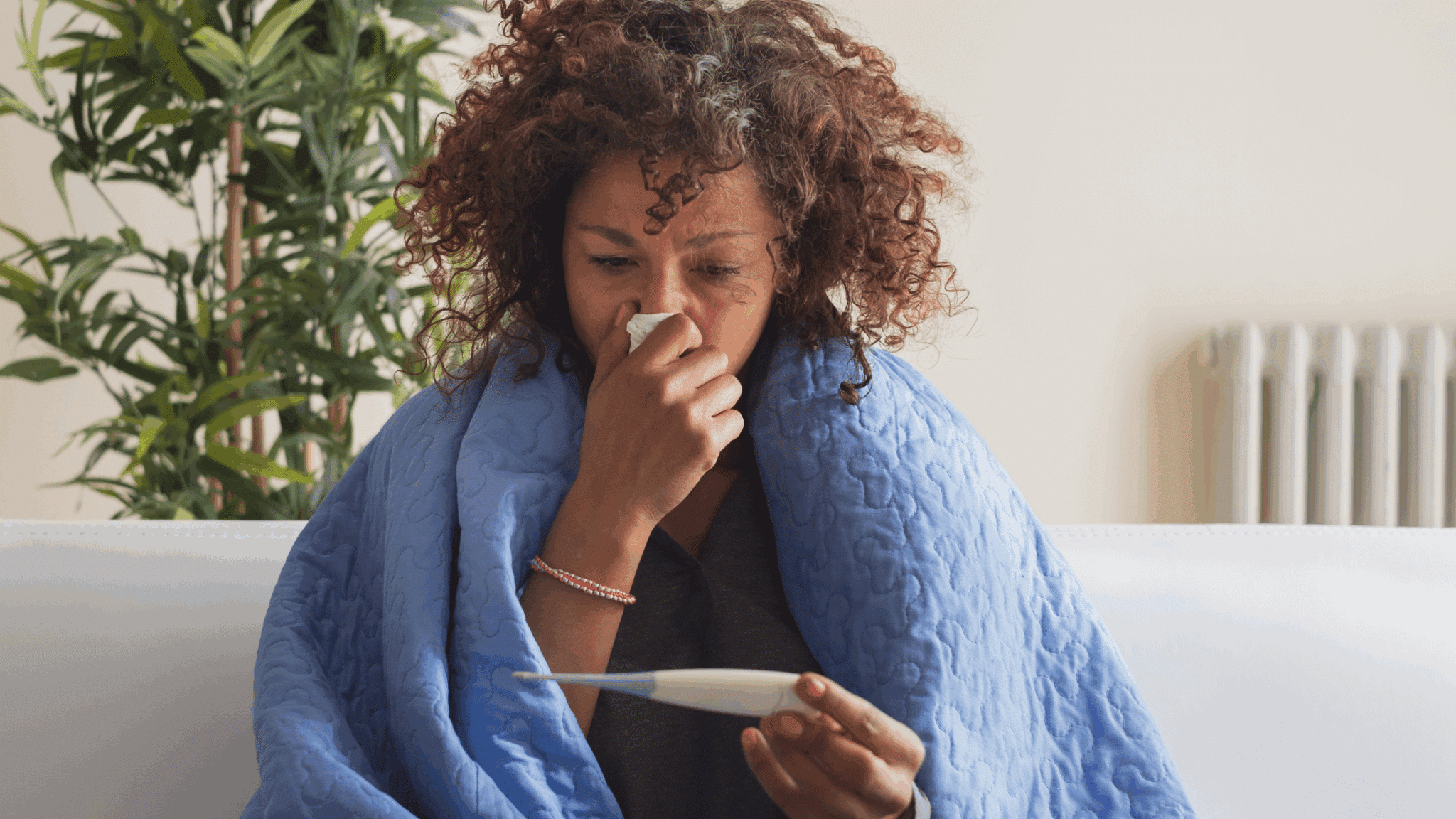 A woman with cold symptoms is checking the thermometer under a blanket.