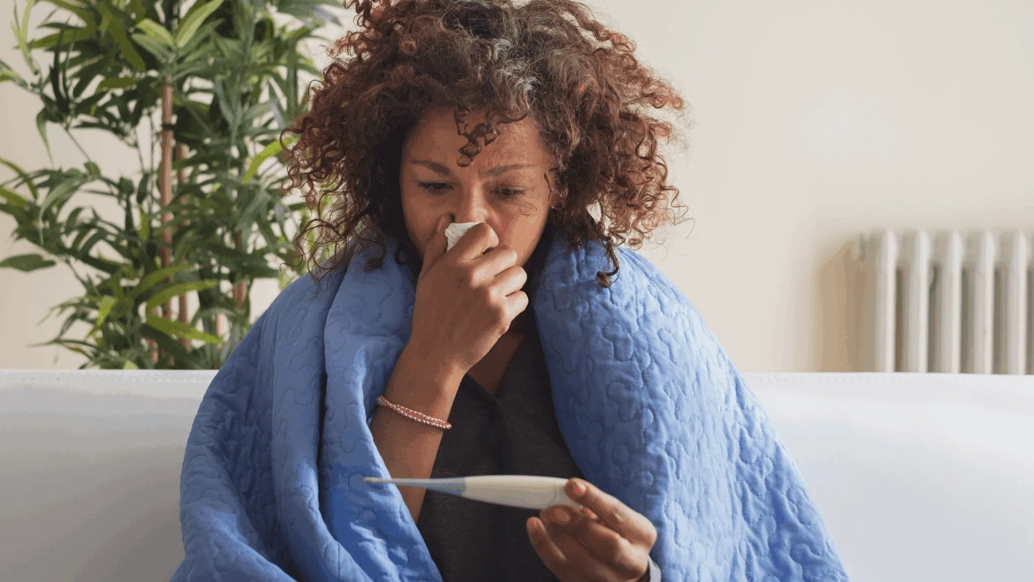 A woman with cold symptoms is checking the thermometer under a blanket.