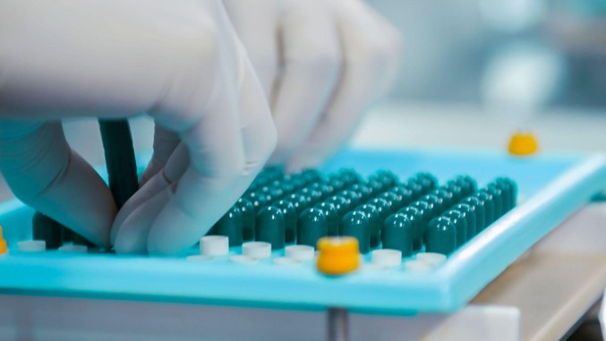 A gloved hand places a small medication vial into a tray of similar capped vials