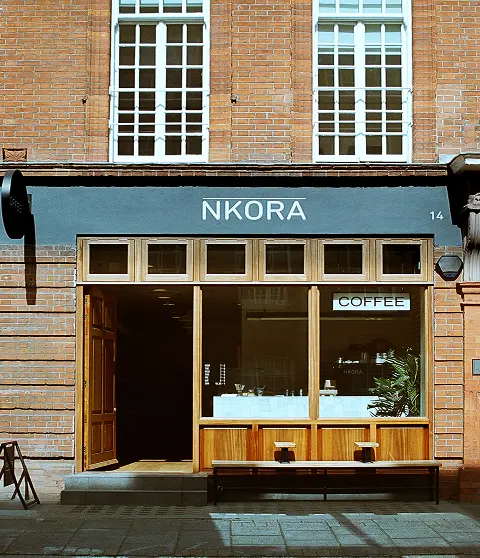 Exterior of NKORA coffee shop with a minimalist wooden storefront, large windows, and a bench outside on a sunny street.