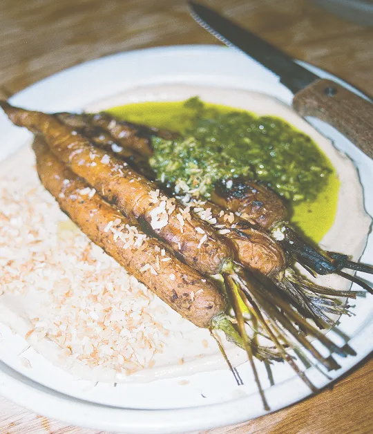 Plate of roasted carrots topped with shredded coconut, served with green herb sauce and a side of rice.