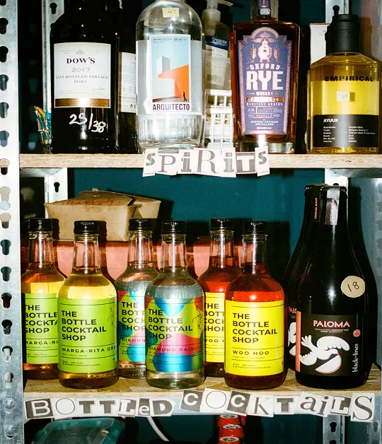 Shelf display of spirits and bottled cocktails, including brands like Oxford Rye, Arquitecto, and The Bottle Cocktail Shop, with handwritten “spirits” and “bottled cocktails” labels.