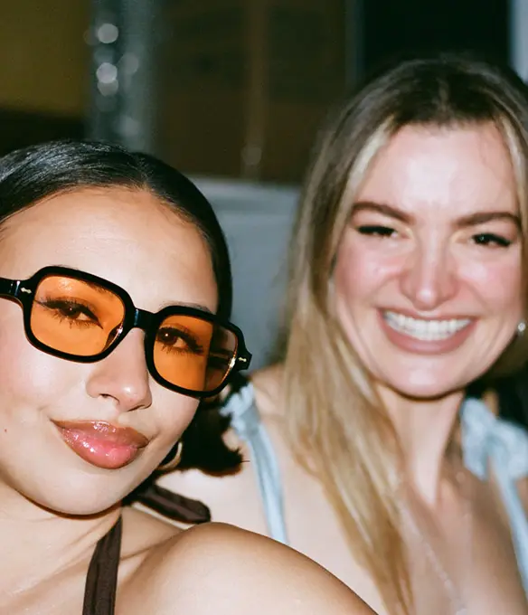 Two women smiling at the camera, one wearing oversized orange-tinted sunglasses, both dressed for a night out.