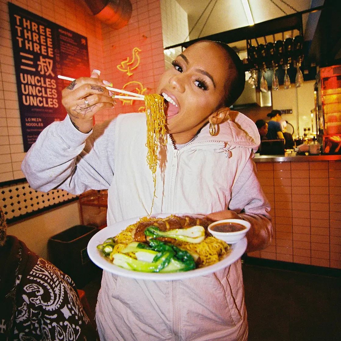 Woman in a white hoodie eating noodles with chopsticks and holding a plate of stir-fried noodles, bok choy, and sauce in a lively restaurant.