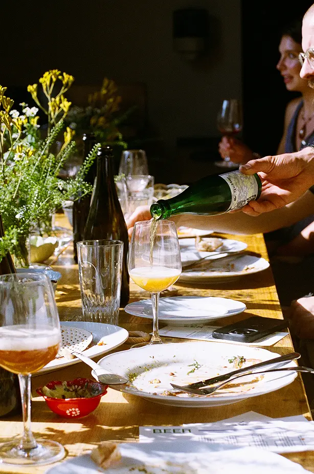 Sunlit outdoor table with empty plates, wine glasses, and wildflowers, as someone pours a drink into a glass during a relaxed group meal.
