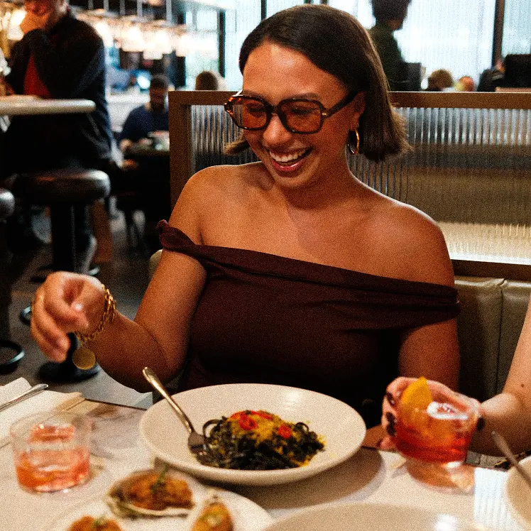 Smiling woman in a burgundy off-shoulder dress at a restaurant table, about to enjoy squid ink pasta with a cocktail in hand.