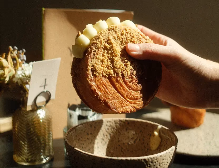 Hand holding a round croissant pastry topped with cream and crumble, over a speckled bowl on a sunlit cafƩ table.