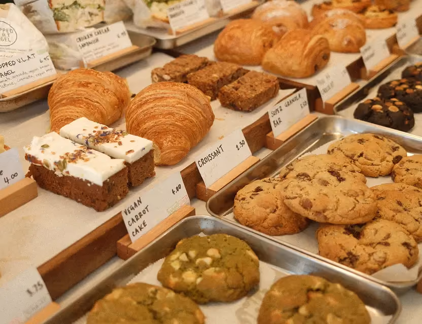 Bakery display with croissants, pain au chocolat, vegan carrot cake, granola bars, and assorted cookies including matcha and chocolate chip.