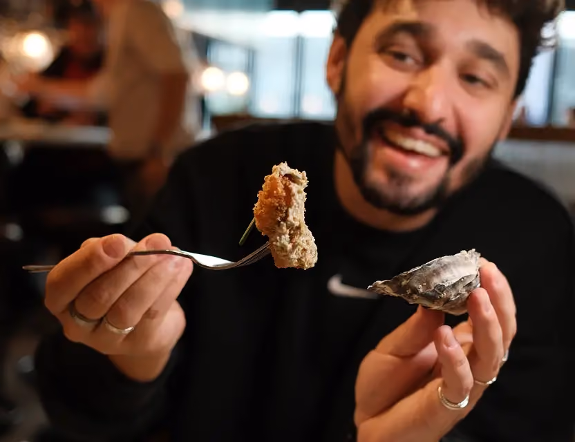 Smiling man holding an oyster shell in one hand and a bite of food on a fork in the other, in a lively restaurant setting.