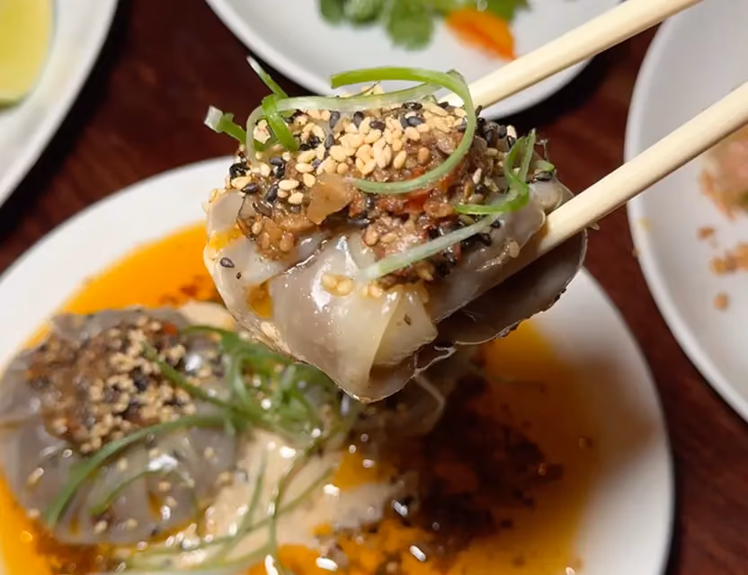 Close-up of a dumpling topped with sesame seeds, scallions, and chili oil, held with chopsticks over a dipping sauce.