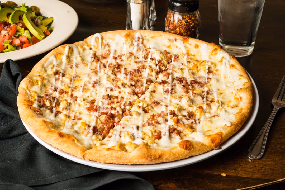 Chicken bacon ranch pizza with creamy white sauce on a white plate with salad and drink in background.