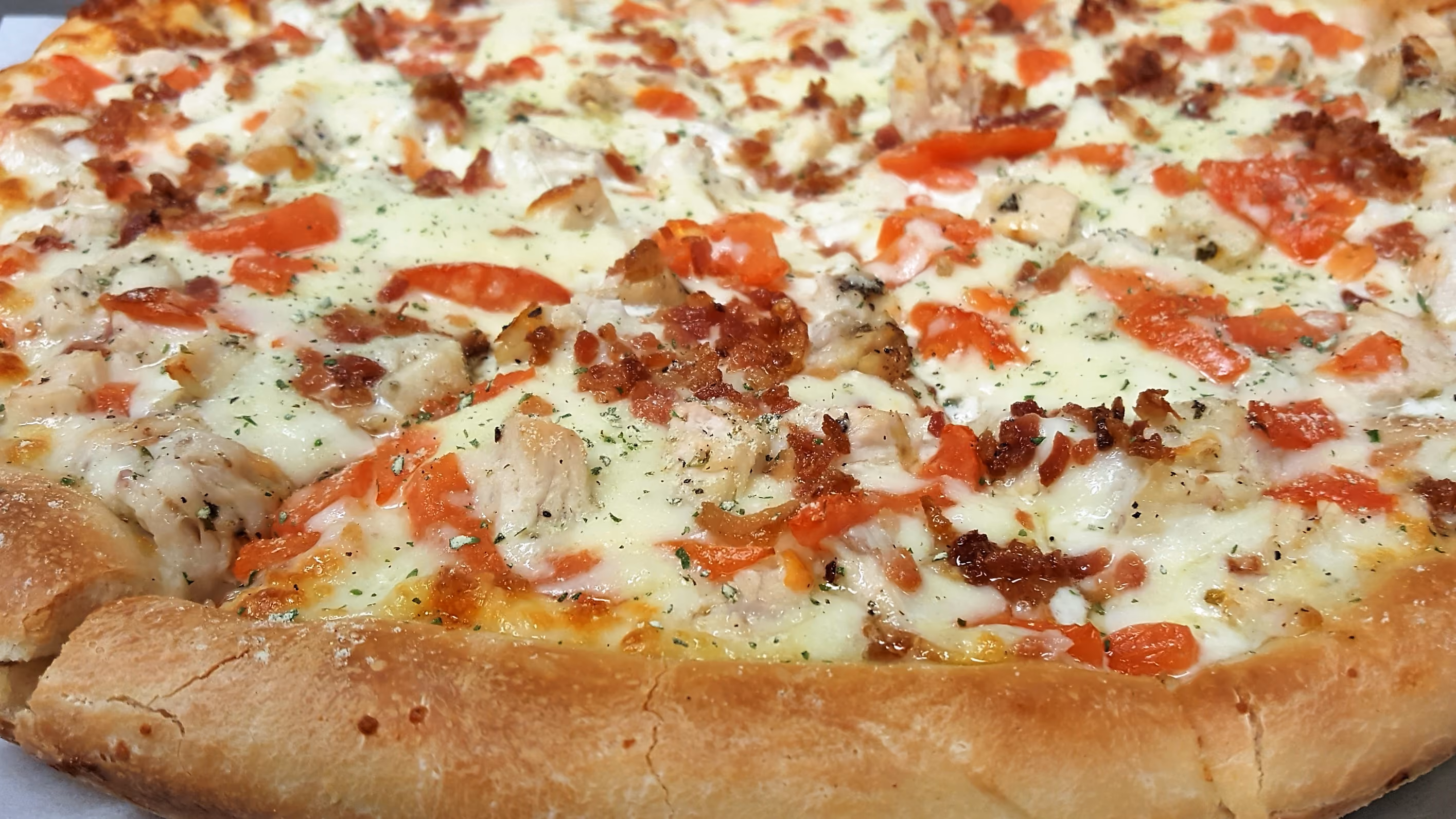 Close-up of pizza with chicken, diced tomatoes, bacon bits, melted cheese, and herbs on crust.