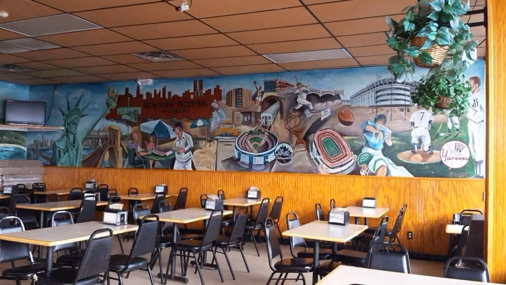 Empty restaurant dining area with tables, chairs, and a mural featuring New York landmarks and sports scenes.