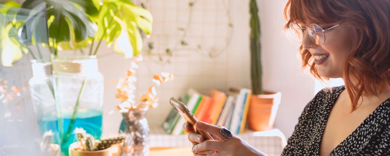 A woman with red hair smiles while using a smartphone in a bright and cozy home office.