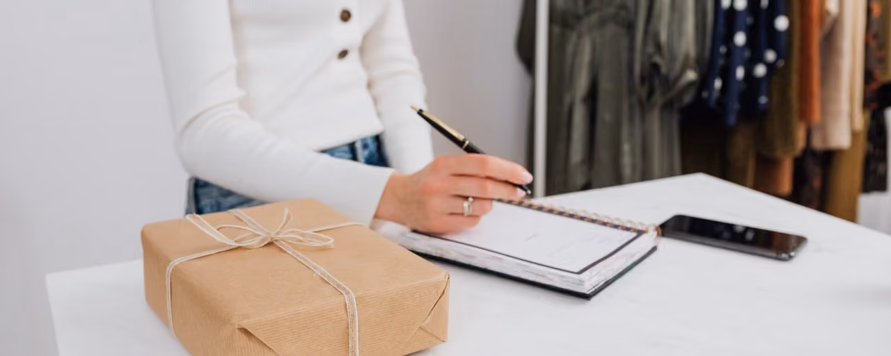 Woman writing in notebook beside a wrapped gift box on a white table indoors.
