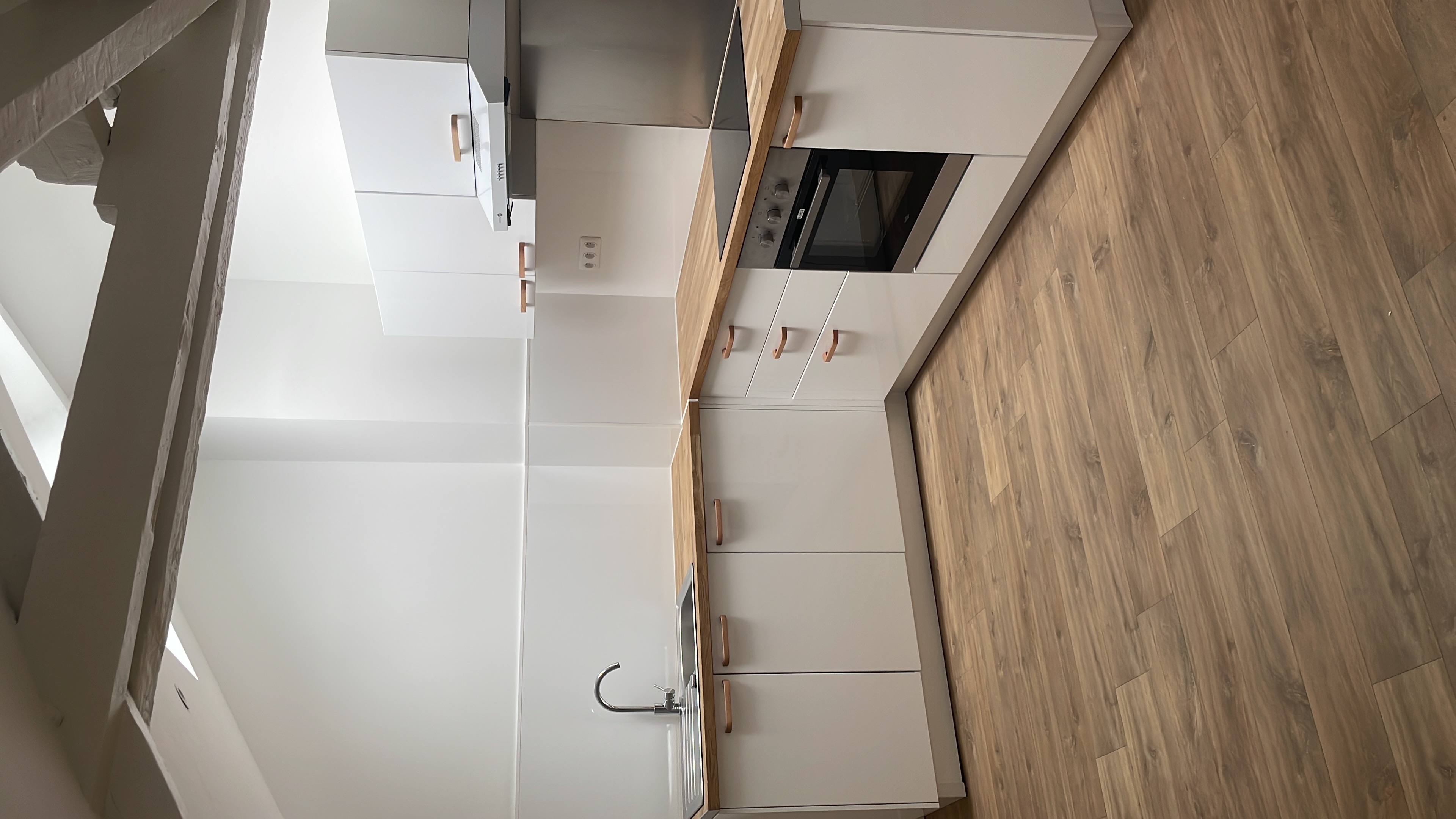Minimalist white kitchen with wooden countertops, built-in oven, sink, and wood floor under angled ceiling beams.