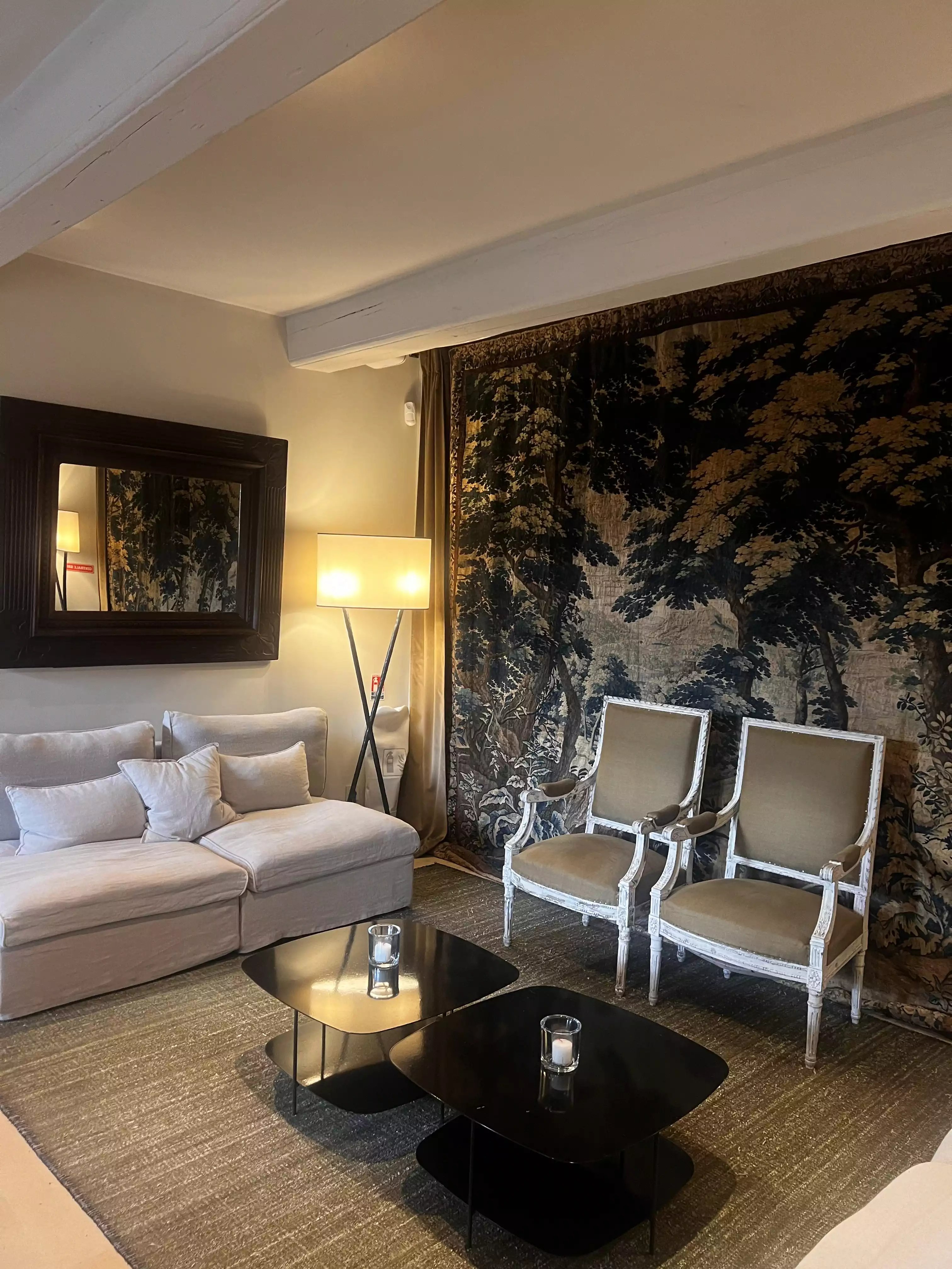 Cozy living room with beige sofa, two armchairs, black coffee tables, tapestry, and floor lamp.
