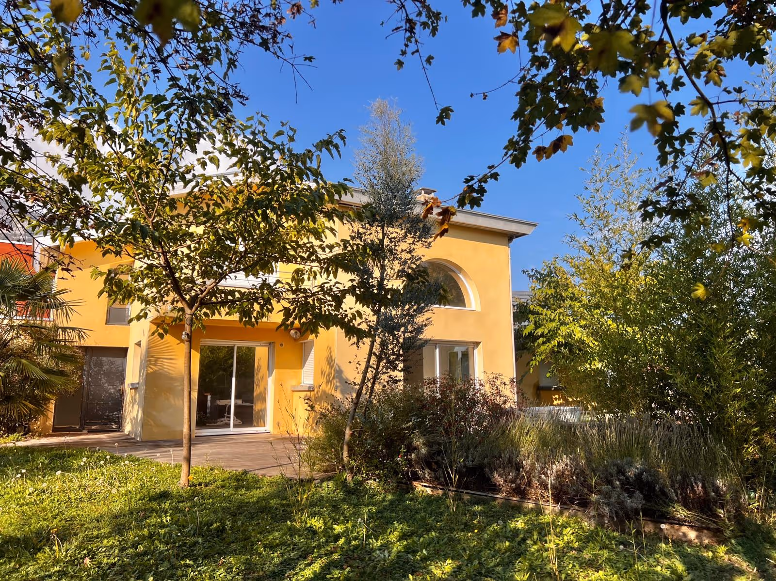 Yellow house with arched and sliding glass doors surrounded by trees and green garden on a sunny day.