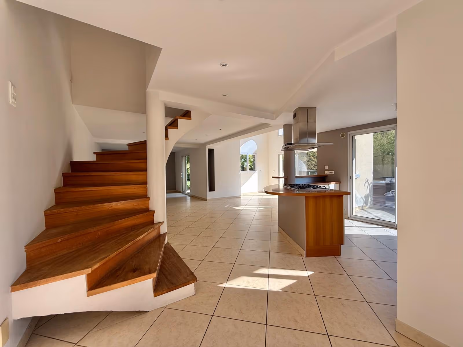Bright open interior with wooden staircase, tiled floor, and kitchen island with stove and vent.