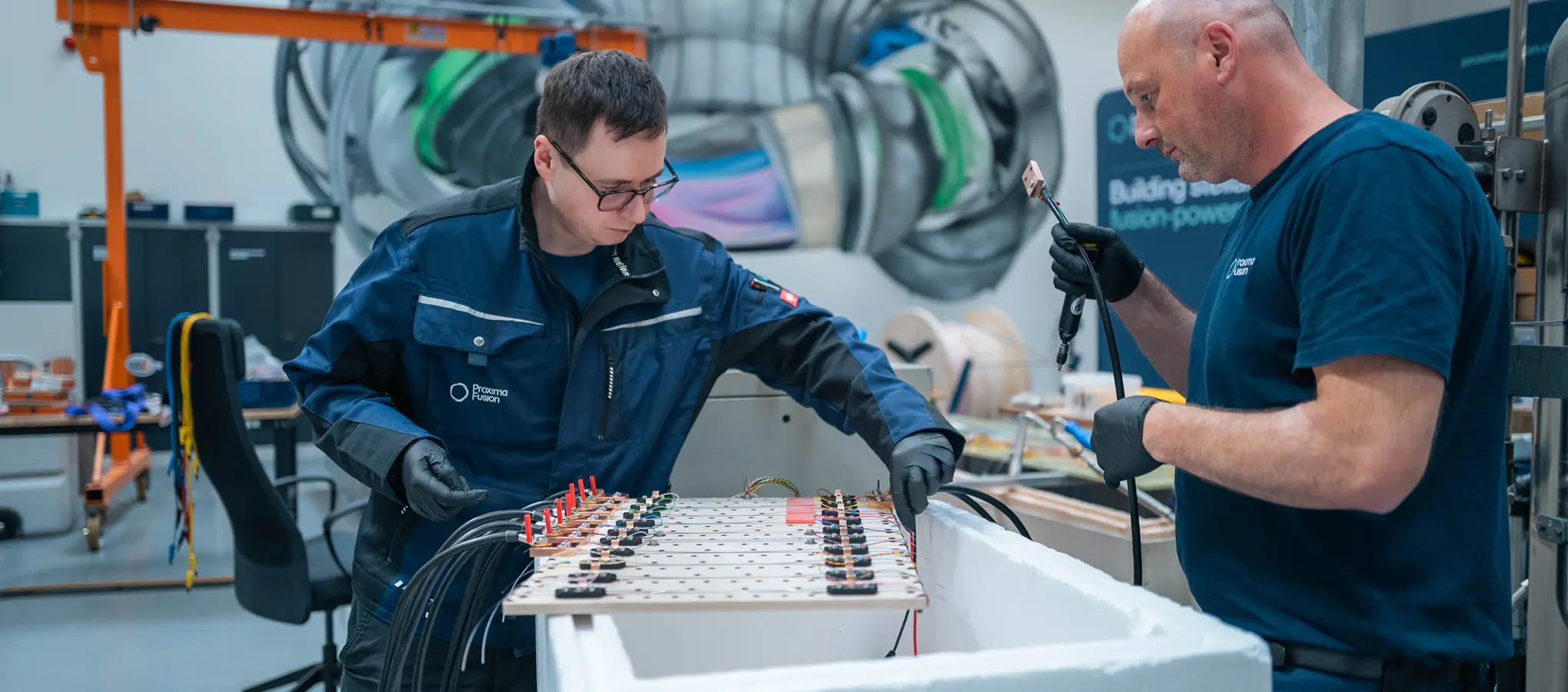 Zwei Proxima-Mitarbeiter arbeiten im Proxima eignen Labor an einem Bauteil mit zahlreichen Kabelanschlüssen um Hochtemperatursupraleiter (HTS) zu testen.