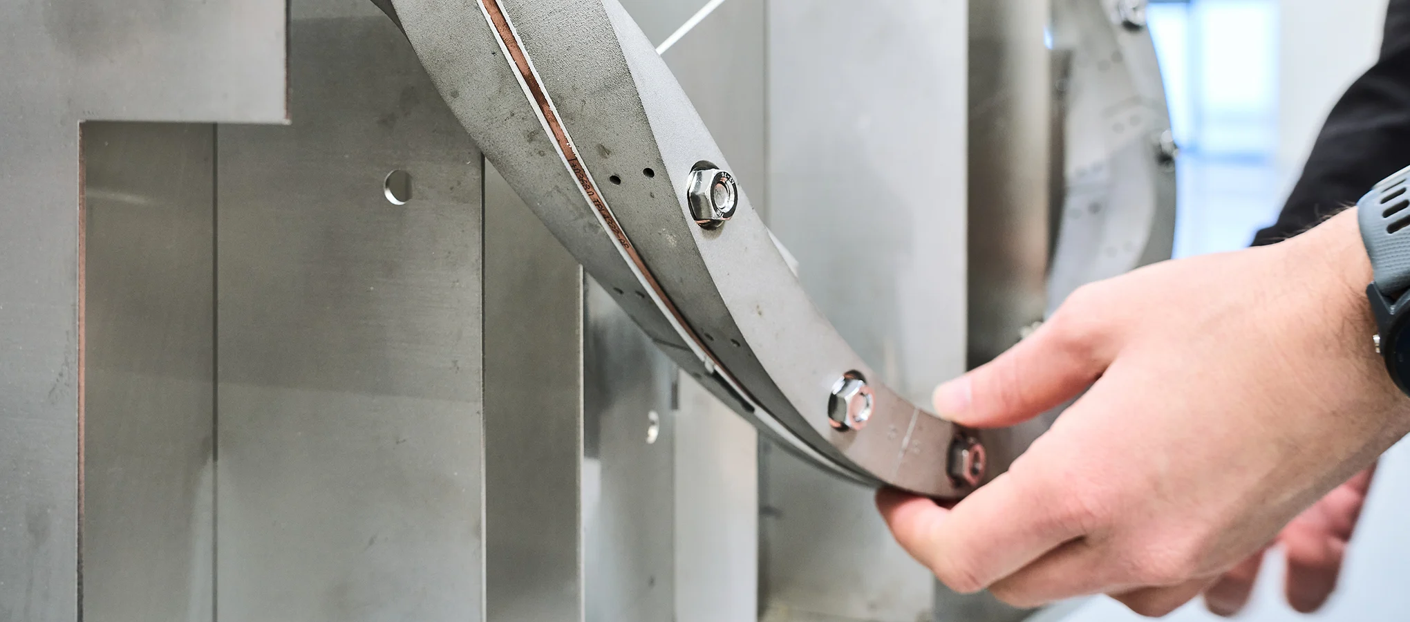 Nahaufnahme einer Hand, die ein gebogenes Metallbauteil mit Schraubverbindungen an einer größeren Konstruktion hält.