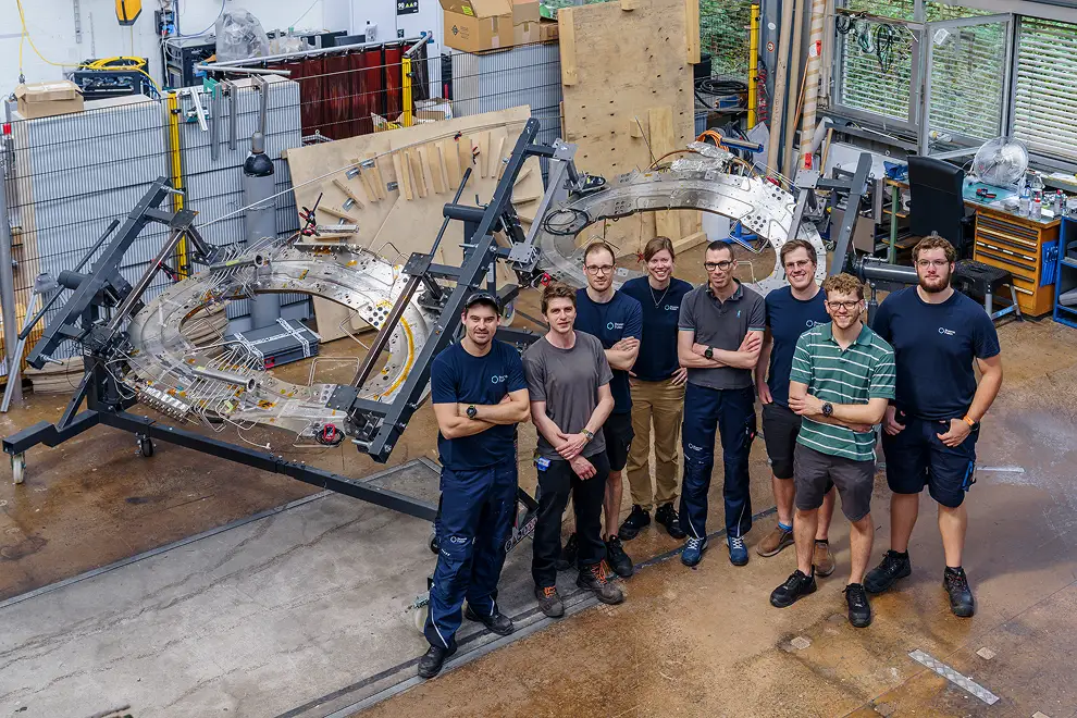 Sieben Mitglieder des Proxima-Fusion-Teams stehen im Labor in Villigen vor ProtoSMC. Hinter ihnen ist der große, ringförmige Versuchsaufbau mit metallischen Halterungen und zahlreichen Kabeln zu sehen.