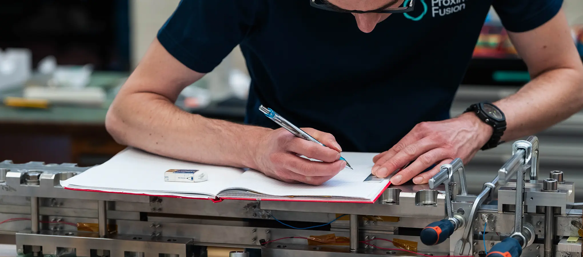 Ein Proxima-Mitarbeiter macht handschriftliche Notizen in einem Heft, das auf einem technischen Versuchsaufbau liegt.
