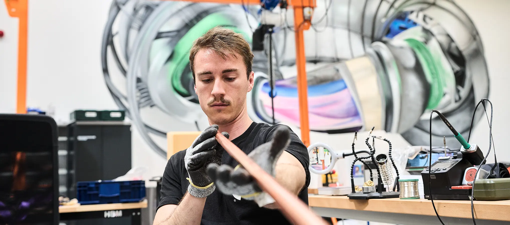 Ein Proxima-Mitarbeiter hält an einem Laborarbeitsplatz ein kupferummanteltes Kabel in behandschuhten Händen. Im Hintergrund ist eine Stellarator-Visualisierung zu sehen.