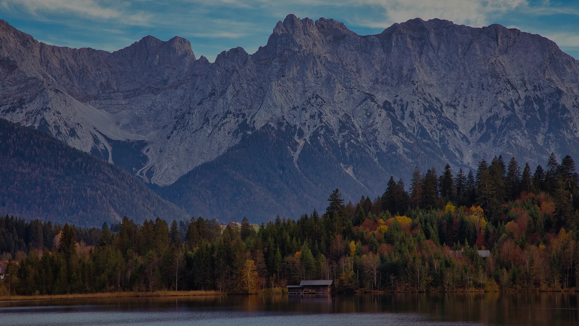 See vor einem herbstlich gefärbten Wald; dahinter erheben sich schroffe Berge in Bayern. 