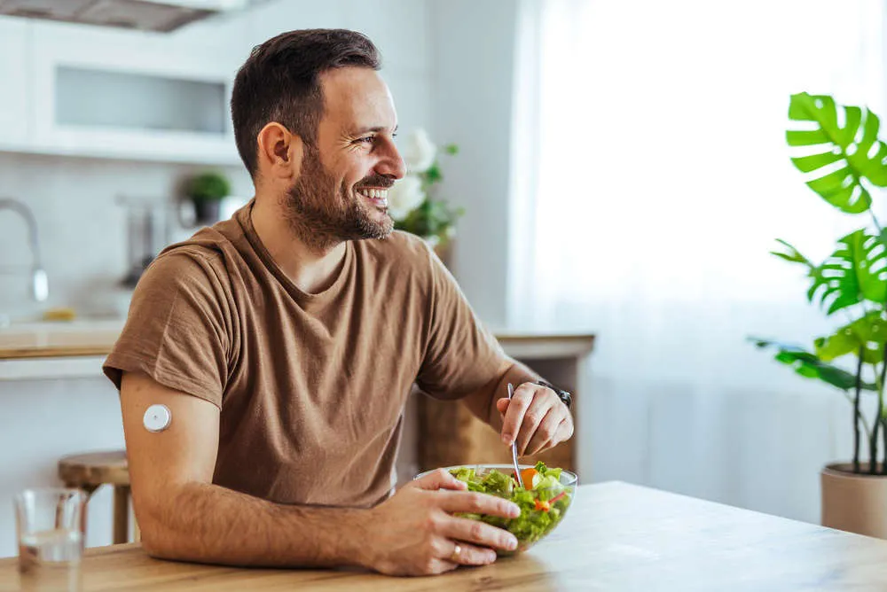 Smiling man wearing a glucose monitor eating a fresh salad at home