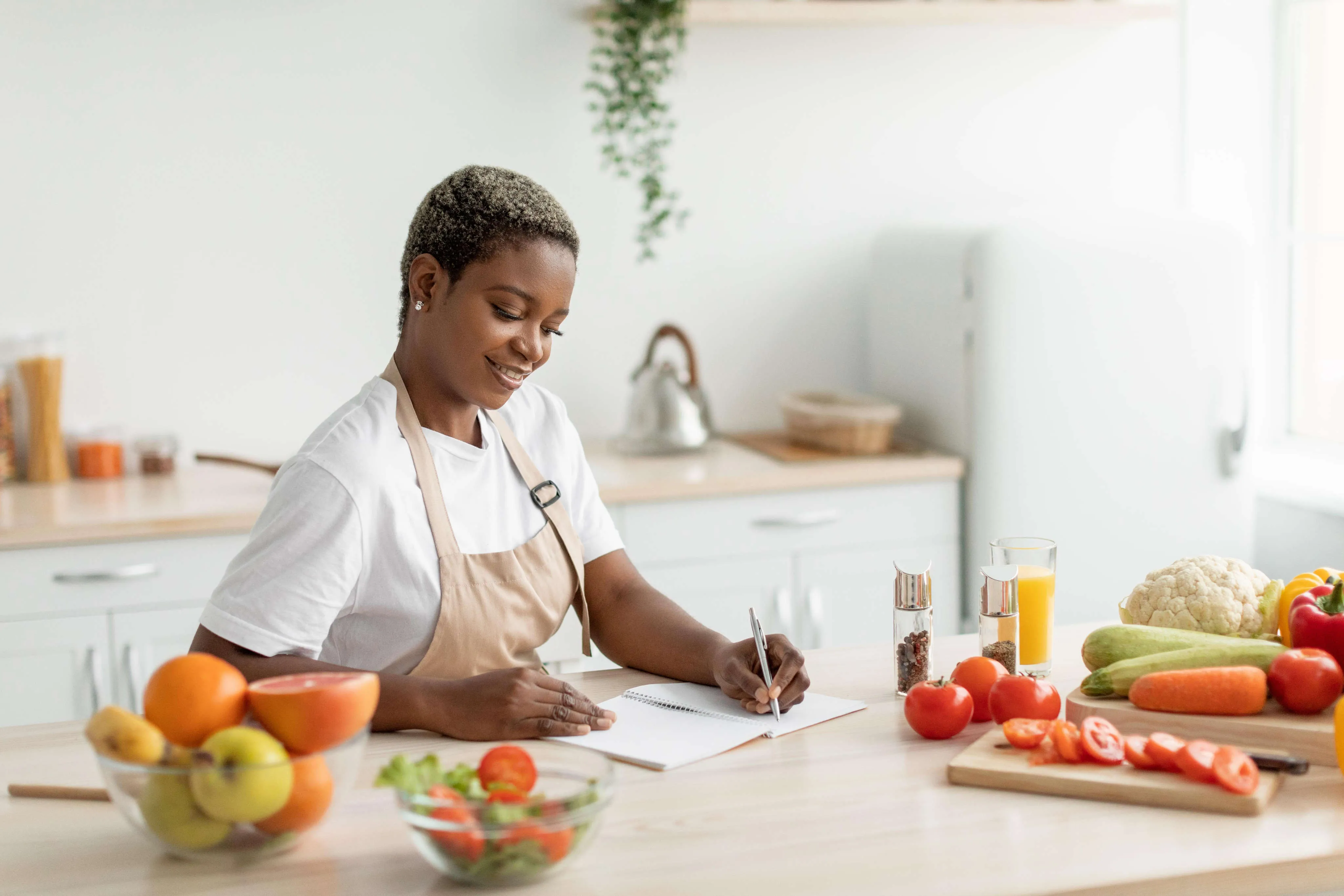 Woman writing a meal plan at her kitchen bench beside fresh fruit and vegetables