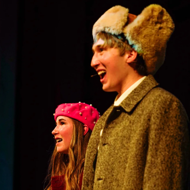 Young man and woman wearing vintage clothing and hats, smiling on stage with dark background.