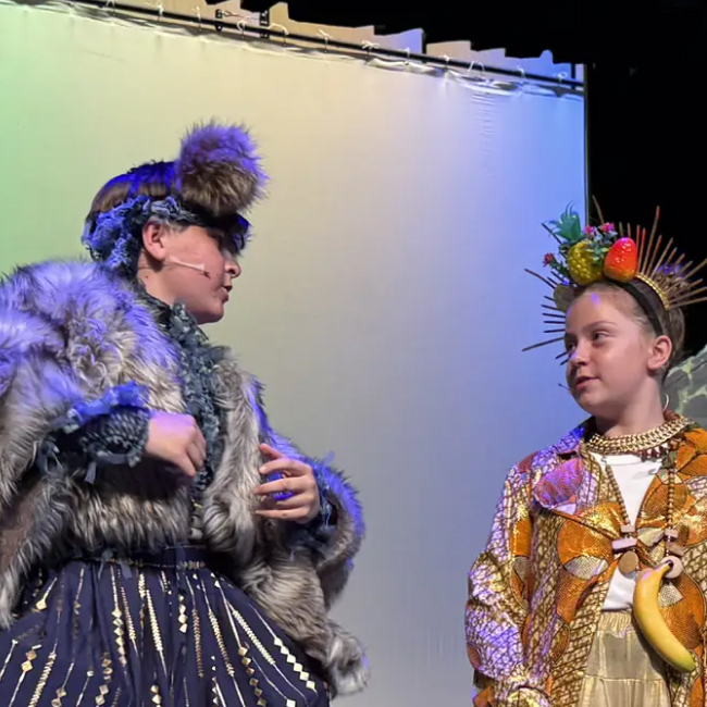 Two children in colorful costumes with headpieces perform on stage against plain backdrop.