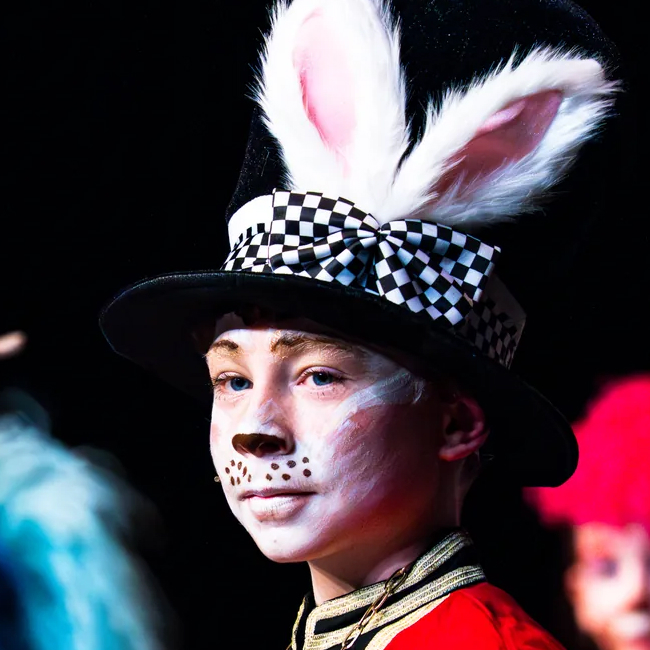 Person wearing bunny ears, checkerboard ribbon hat, and face paint resembling a rabbit.