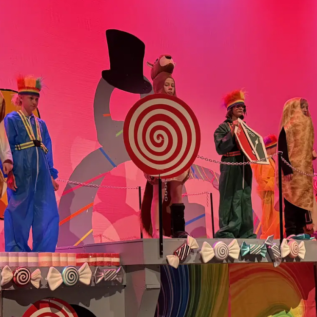 Children in colorful costumes on a stage with candy and target props under pink lighting.