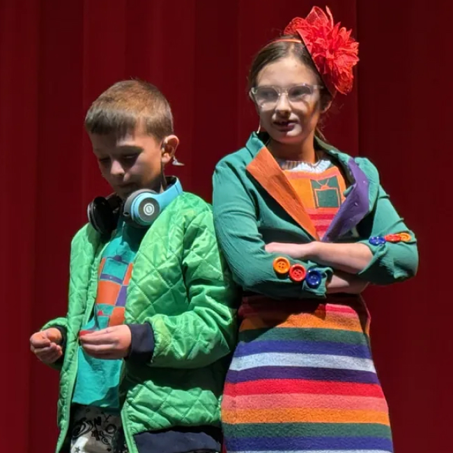 Two children in colorful clothes standing on stage against a red curtain backdrop.