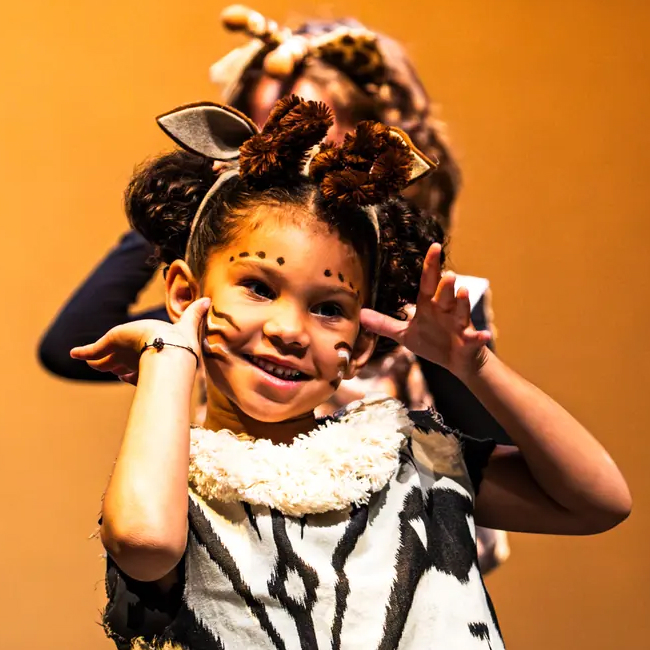 Smiling child in animal costume with painted whiskers making paws gesture on orange background.