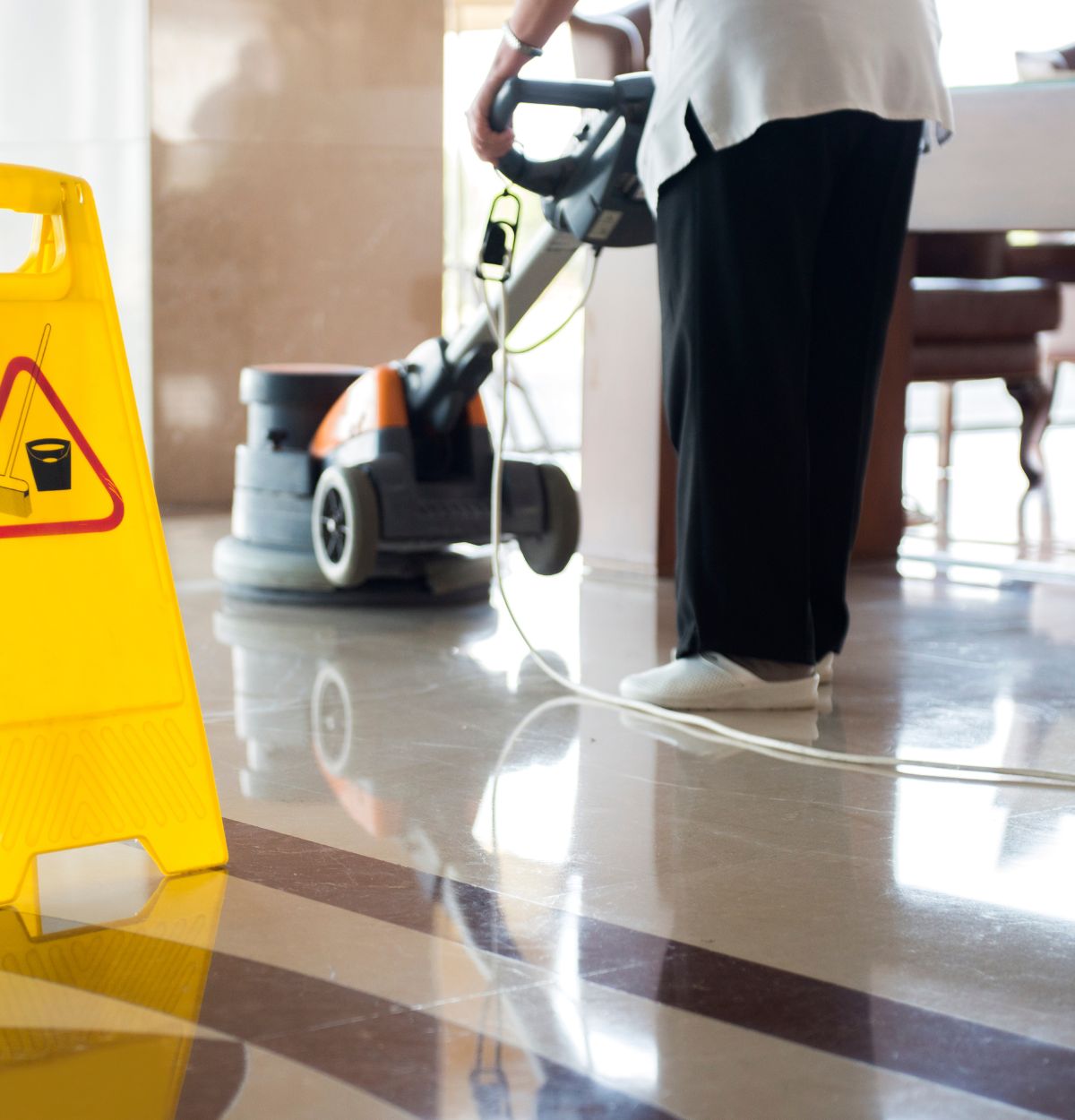 Person operating an electric floor cleaning machine on a shiny tiled floor with a yellow caution sign in the foreground.