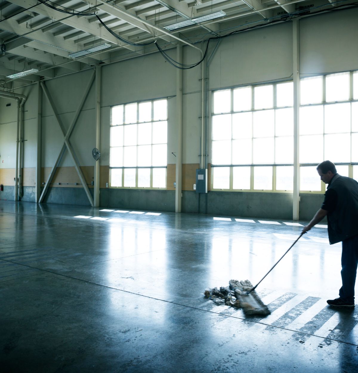 A person sweeping a polished concrete floor in a large, empty industrial warehouse with high ceilings and large windows letting in bright natural light.